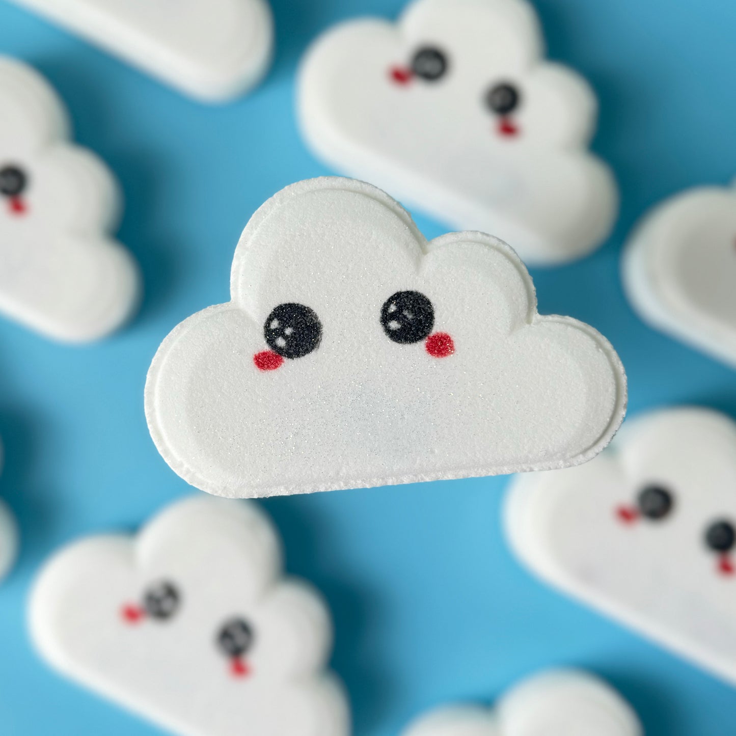 Close up of a cloud shaped bath bomb, with cute eyes and rosey cheeks, on a blue background. 